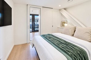 Ein Schlafzimmer mit einem Bett, einer grünen Decke, einer weißen Wand, einer Tür, die auf einen Balkon führt, und einem an der Wand befestigten Fernseher.