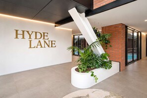 Eine moderne Hotelhalle mit einem weißen Empfangsschalter, einem großen Schild mit der Aufschrift 'HYDE LANE' und einem Blumentopf mit Grünpflanzen.