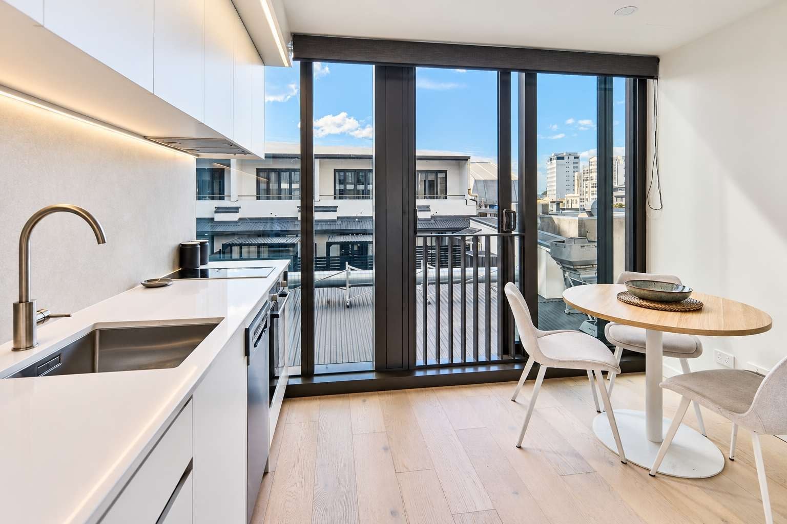 Bright Te Aro apartment kitchen and dining area with seating for three, generous bench space, modern cabinetry and sliding glass doors opening to a compact balcony with an urban outlook.