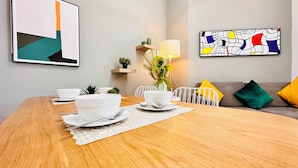 Mesa de jantar posta com tigelas e pratos brancos, um vaso com plantas verdes, uma luminária e um quadro abstrato colorido na parede.