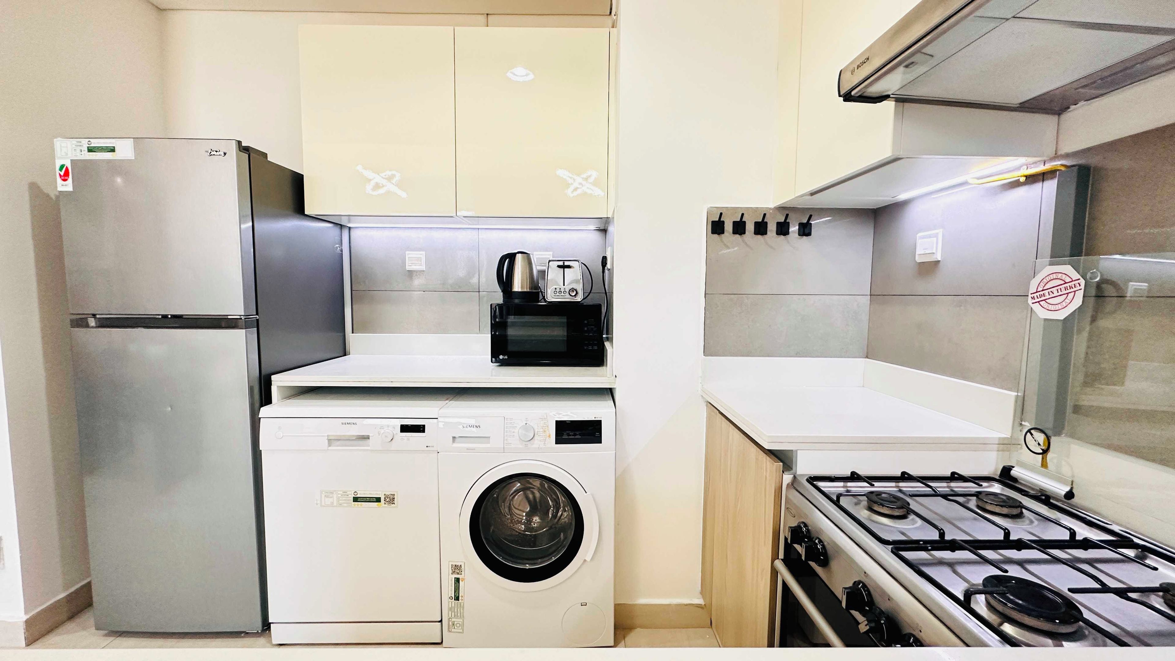 A compact kitchen with a stainless steel refrigerator, a microwave, a washing machine, and a gas stove.
