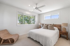 A bedroom with a bed, a ceiling fan, two bedside tables with lamps, a chair, and a window with a view of trees. - Whale Watcher - elevated coastal escape (Lennox Head)