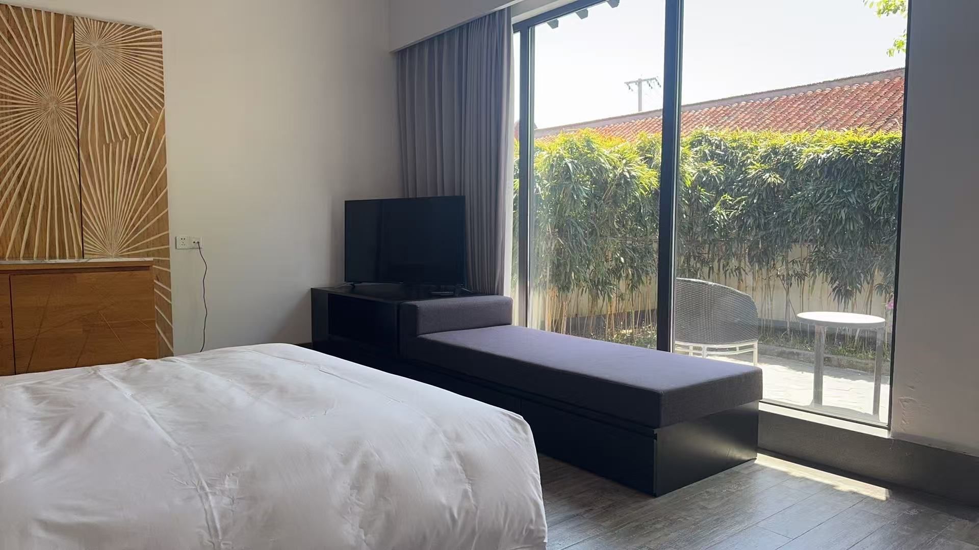 A modern hotel room with a bed, a TV on a stand, and a view of a garden through large windows.