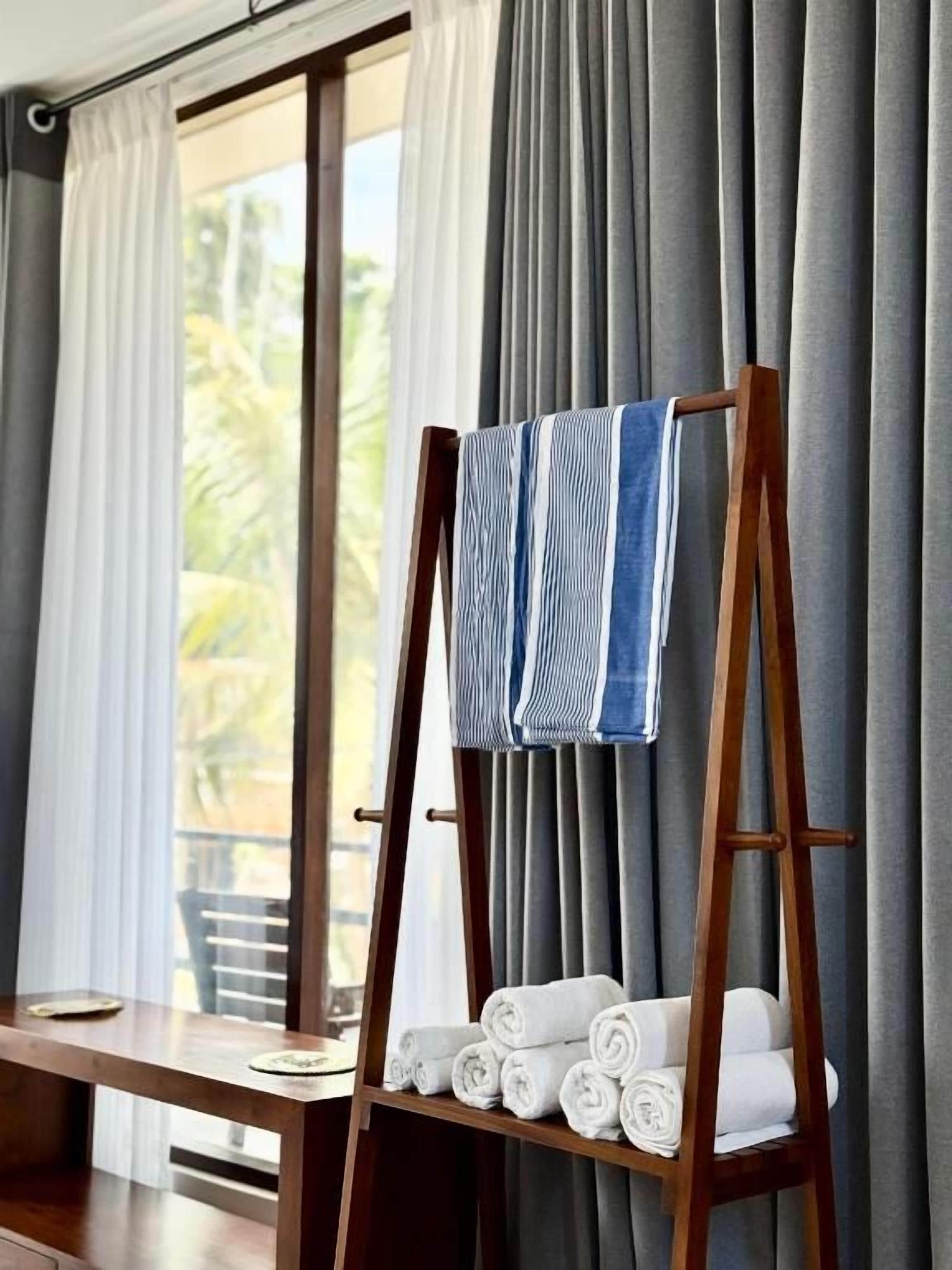 A wooden easel with rolled towels and a blue and white striped towel on it, placed in front of a window with curtains.