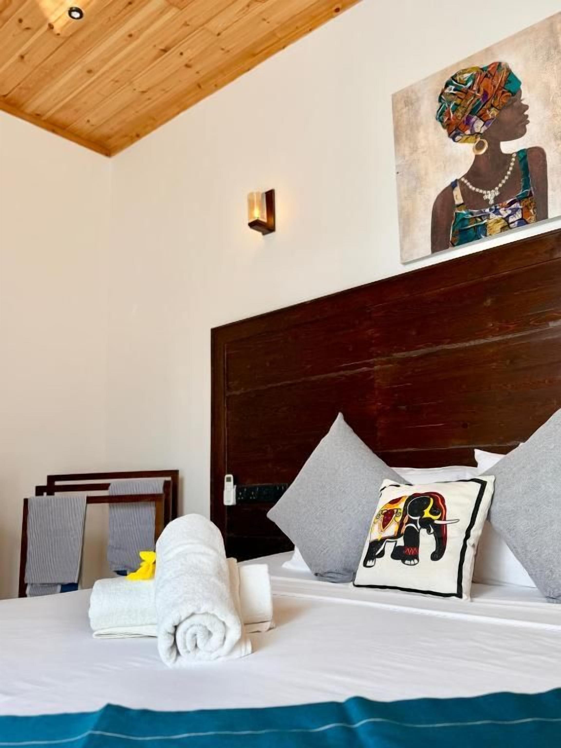 Ein Hotelzimmer mit Holzbalkendecke, einem großen Bett mit Kissen, einem Gemälde über dem Bett und einem kleinen Kissen mit Elefantenmotiv.
