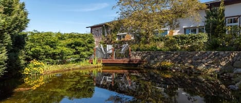 A house with a stone bridge over a pond, surrounded by lush greenery and trees.
