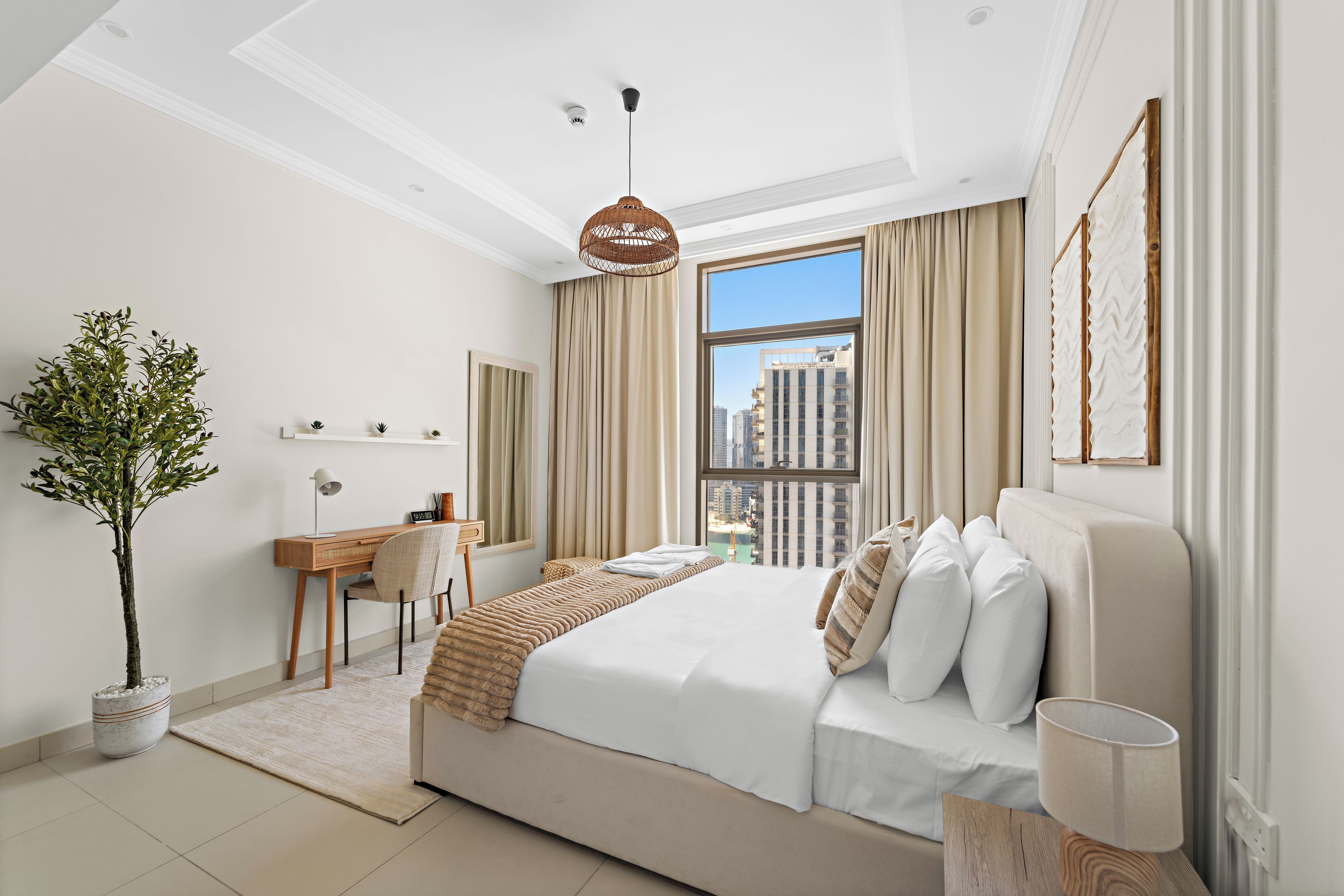 A modern hotel room with a large bed, a desk, a chair, a potted plant, and a view of the cityscape.