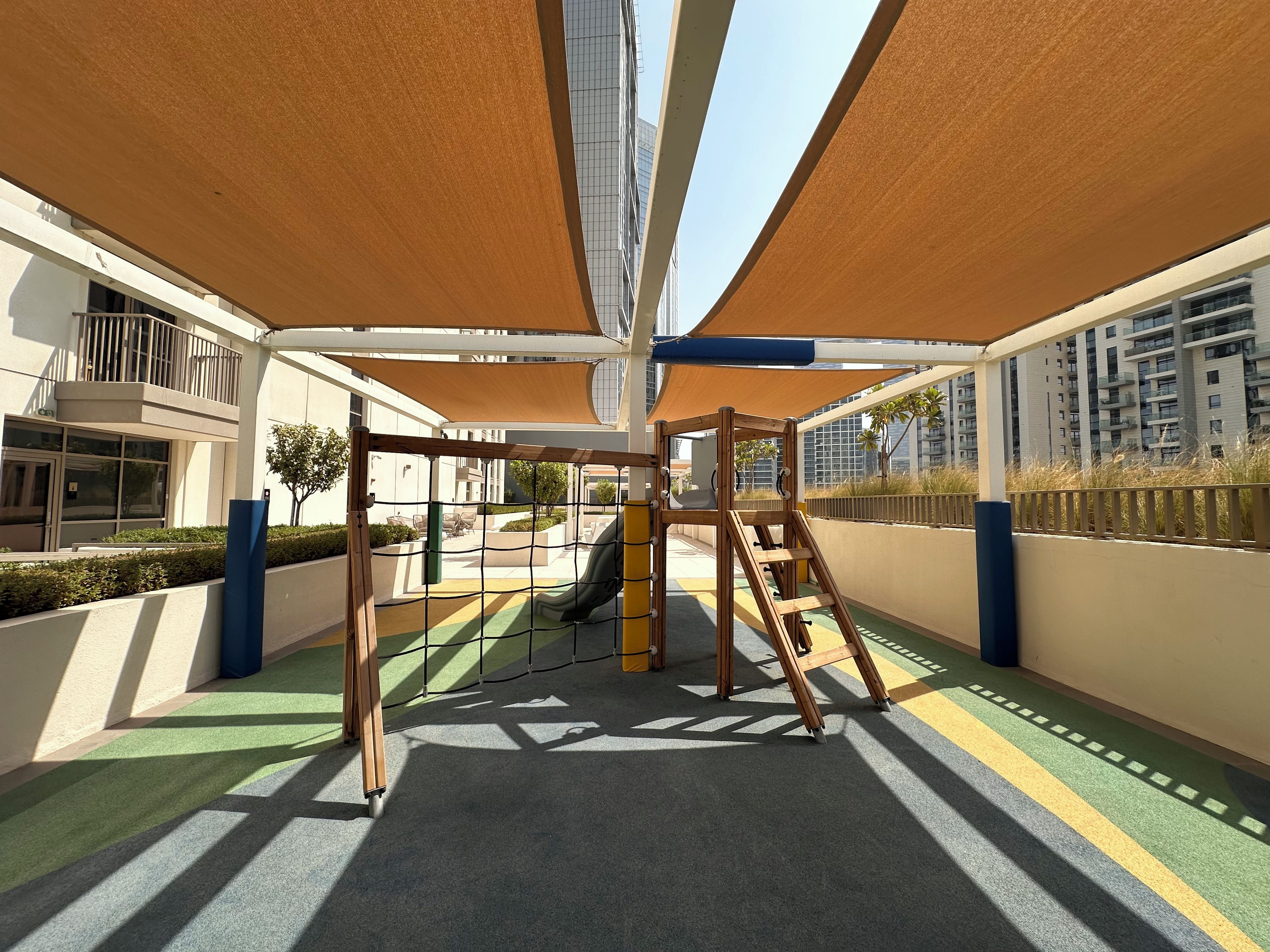 A playground with a slide and climbing structures, surrounded by shaded areas and a view of residential buildings.