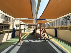 A playground with a slide and climbing structures, surrounded by shaded areas and a view of residential buildings. - Vogue | Reflection near Reem Central Park & Beach (Abu Dhabi)