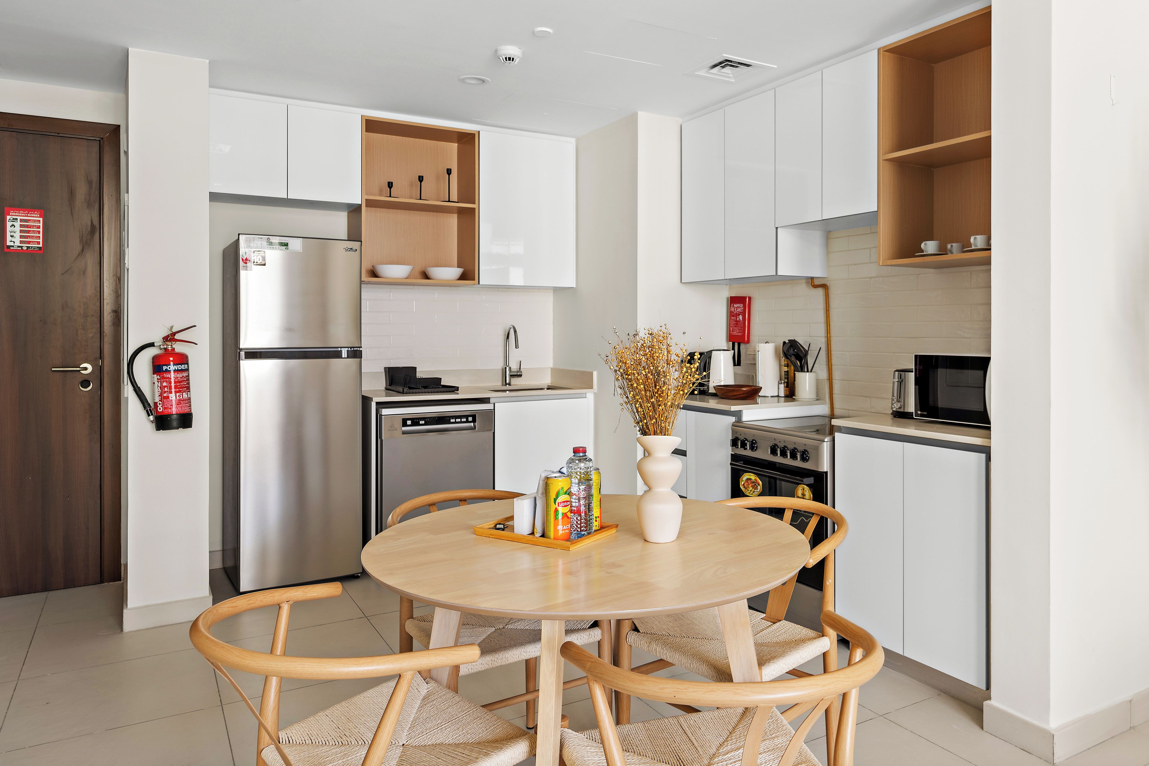 Une cuisine moderne avec des meubles blancs, un réfrigérateur en acier inoxydable et un coin repas comprenant une table ronde et des chaises en osier.