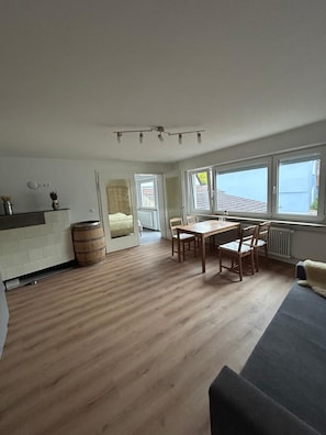 A spacious living area with a wooden floor, a dining table with chairs, a sofa, and a view of the sea through large windows.