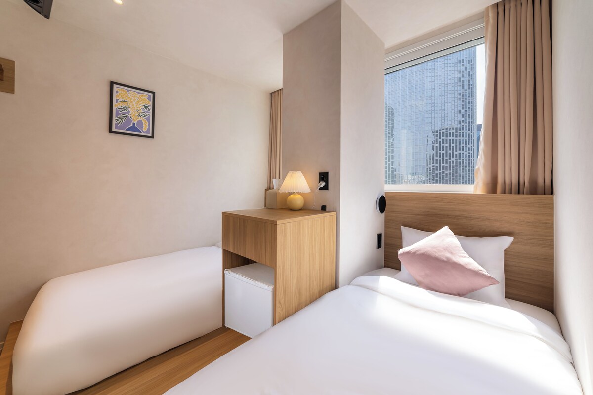 A hotel room with two beds, a nightstand with a lamp, a small refrigerator, a window with curtains, and a framed picture on the wall.