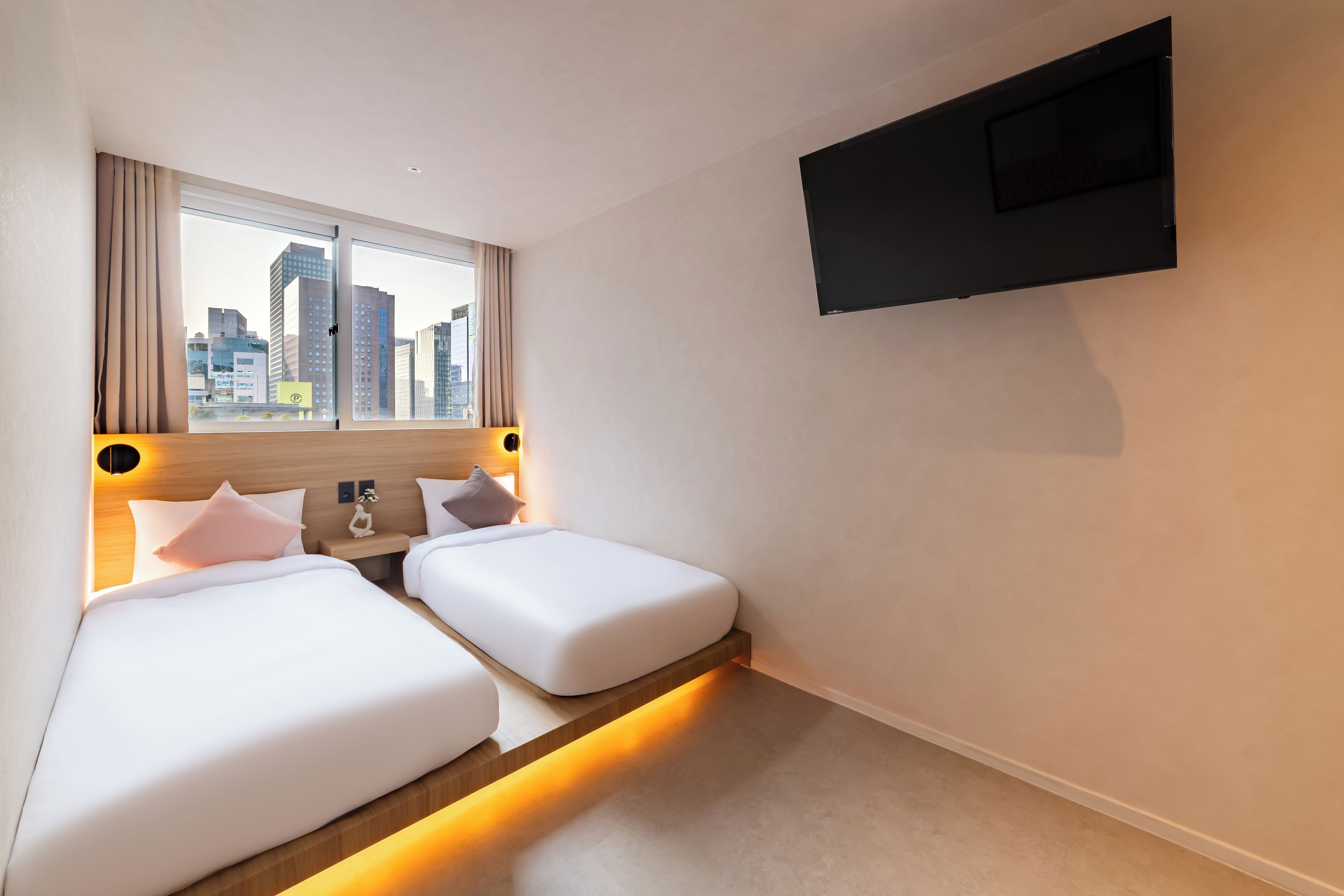 A hotel room with two beds, a city view, a flat-screen TV mounted on the wall, and a small bedside table with a vase.