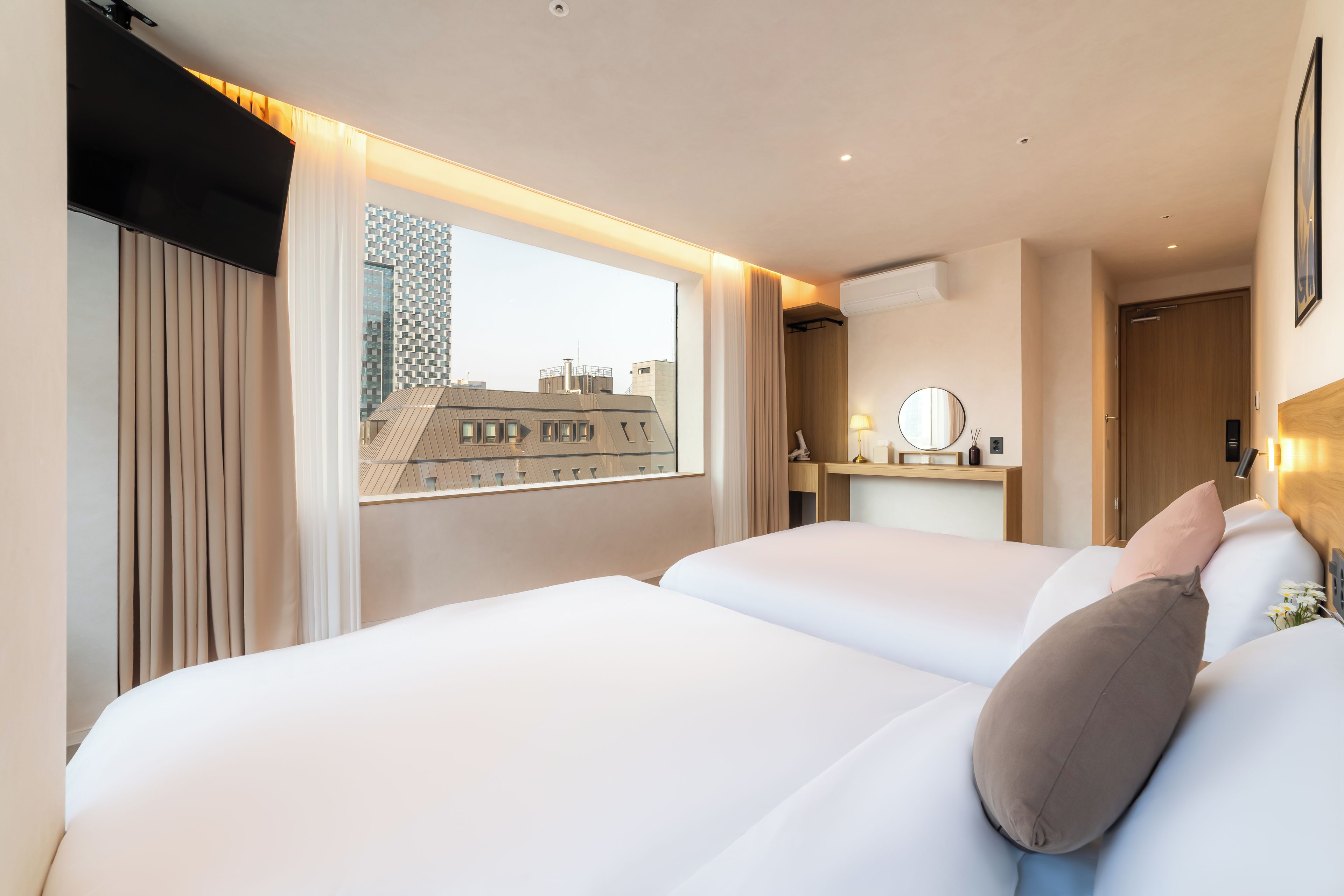 A hotel room with two beds, a window with curtains, a bedside table with a lamp, and a view of a building outside.