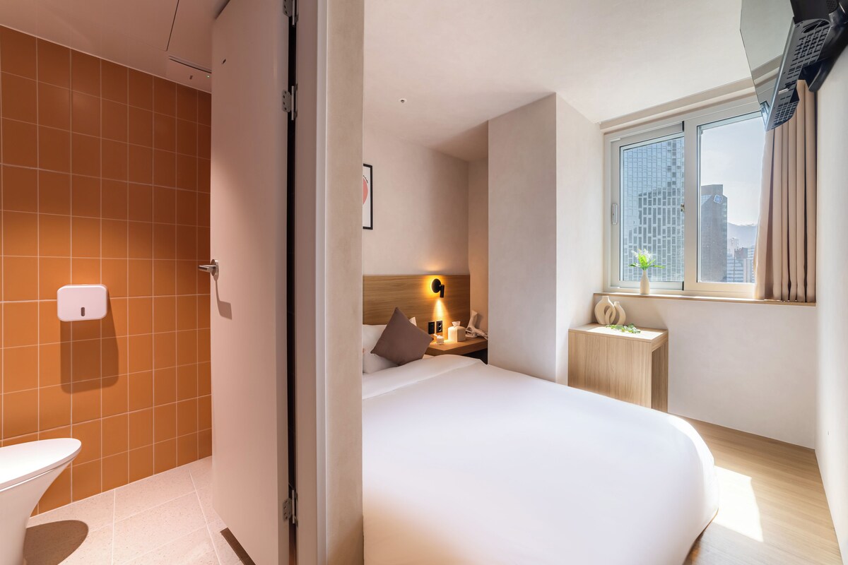 A modern hotel room with a bed, bedside table, and a window with a city view.