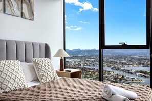 A bedroom with a large window, a bed with a textured blanket, two pillows, a bedside lamp, and a view of a cityscape and mountains. - Stunning Broadbeach 1BR with Sweeping City Views (Gold Coast)
