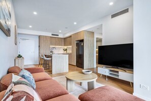 A modern living room with a sofa, a coffee table, and a TV. There is a kitchen area with a fridge and a dining table. - Stunning Broadbeach 1BR with Sweeping City Views (Gold Coast)