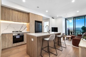 A modern kitchen with a marble backsplash, wooden cabinets, and a central island with bar stools. - Stunning Broadbeach 1BR with Sweeping City Views (Gold Coast)