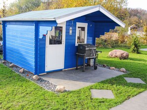 Eine kleine, blaue Gartenhütte mit Tür und Fenstern, einem Grill und einer gepflasterten Fläche mit Gras und Steinen.