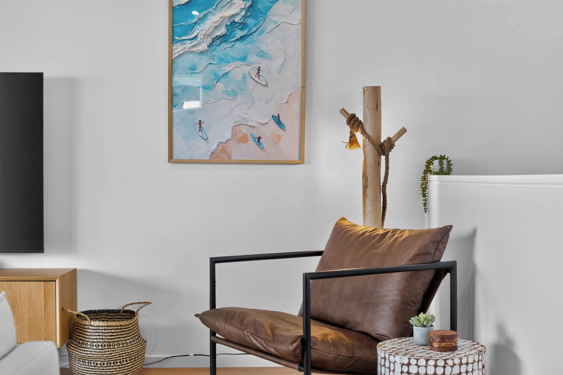 A modern living room with a leather armchair, a woven basket, a small round table with a plant, and a large abstract painting on the wall.