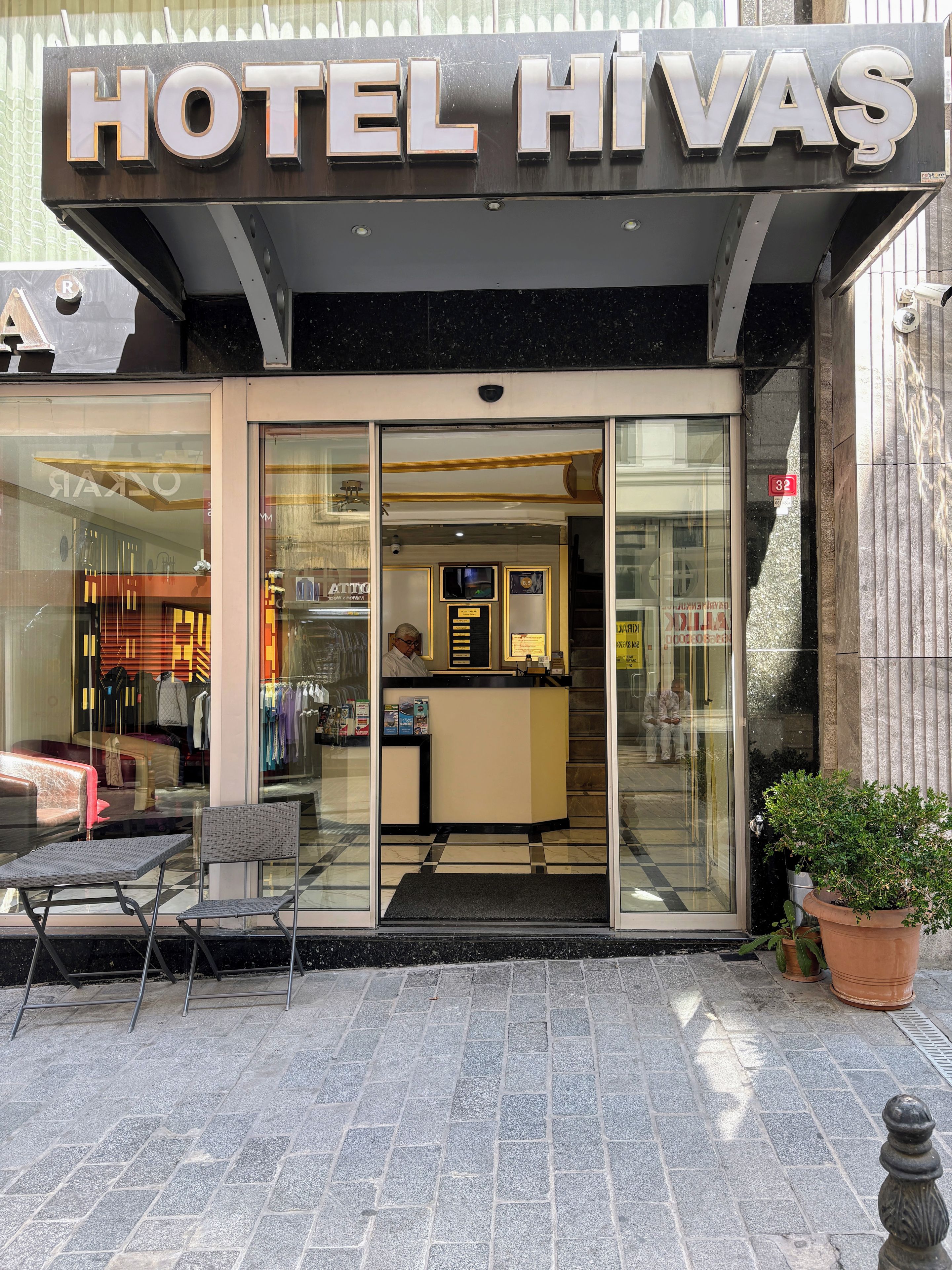 A hotel entrance with a glass door, a reception desk, and a seating area outside.