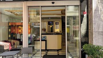 A hotel entrance with a glass door, a reception desk, and a seating area outside.
