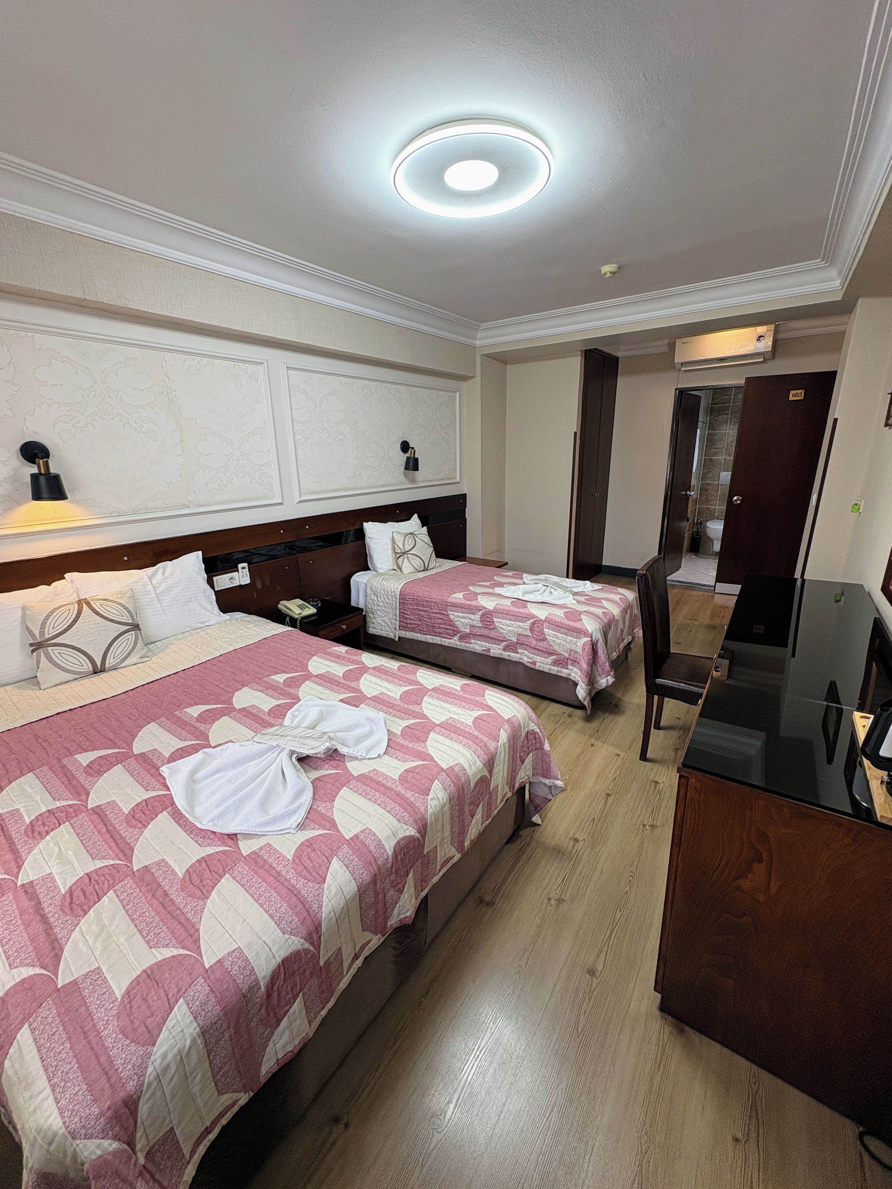 A hotel room with two beds, a desk, a mirror, and a bathroom door.
