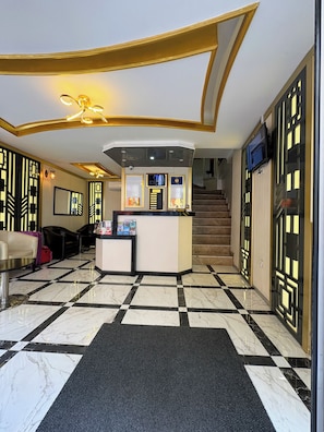 Un hall d’hôtel comprenant un comptoir d’accueil, des guichets d’enregistrement, un escalier et un espace dédié à l’enregistrement des clients.