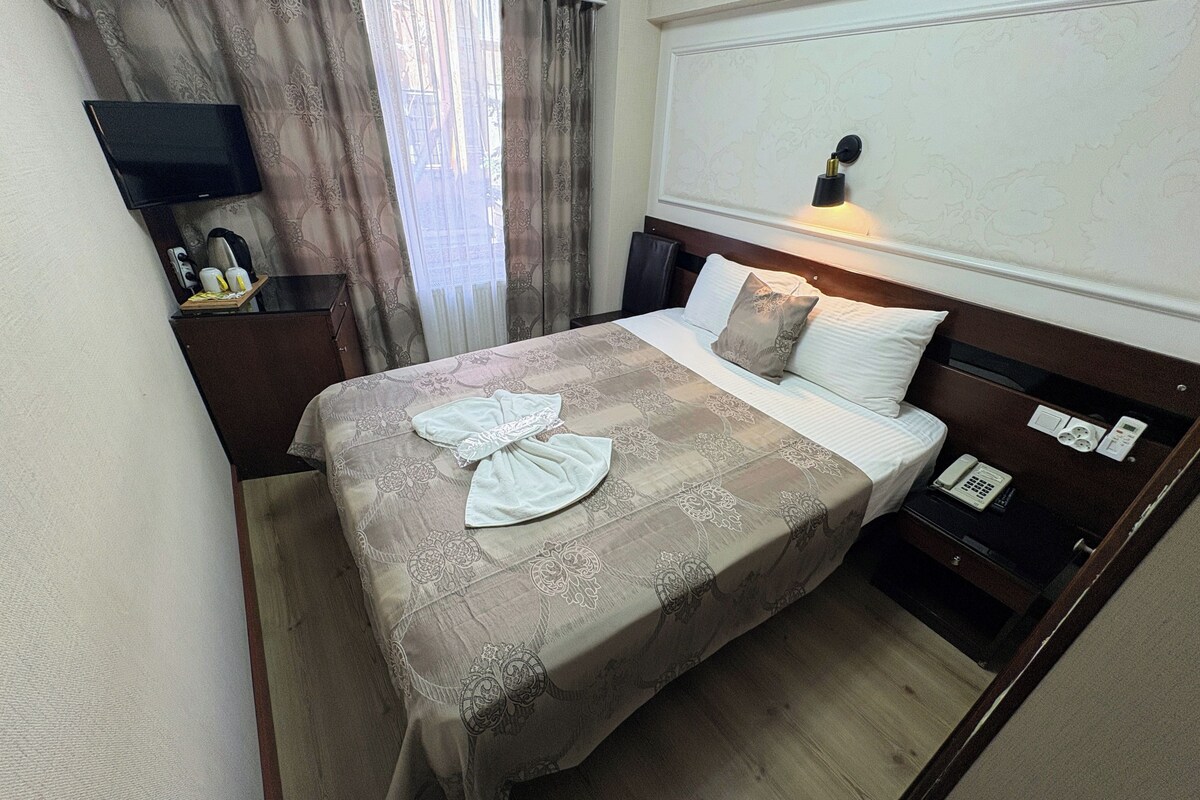 A hotel room with a bed, a bedside table with a phone, a wall-mounted light, and a television on a stand.