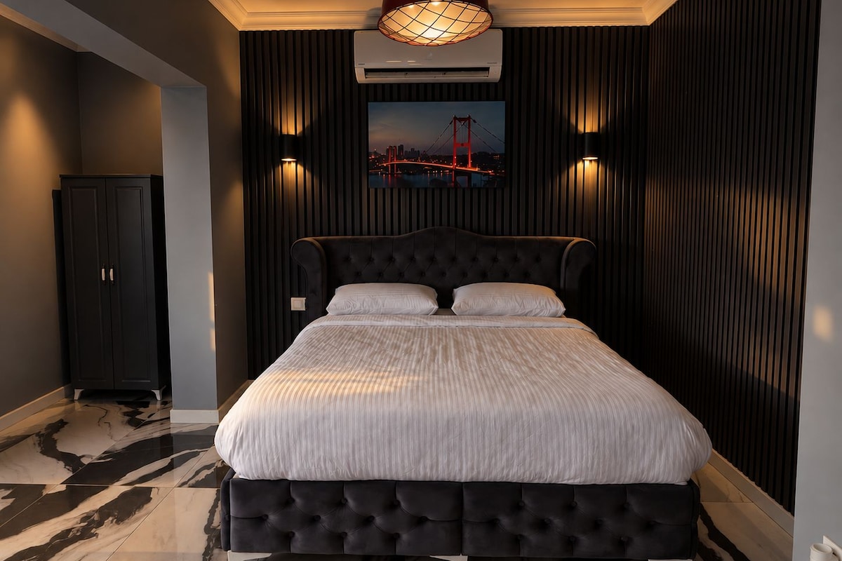 A bedroom with a large bed, dark walls, and a cityscape painting above the bed.