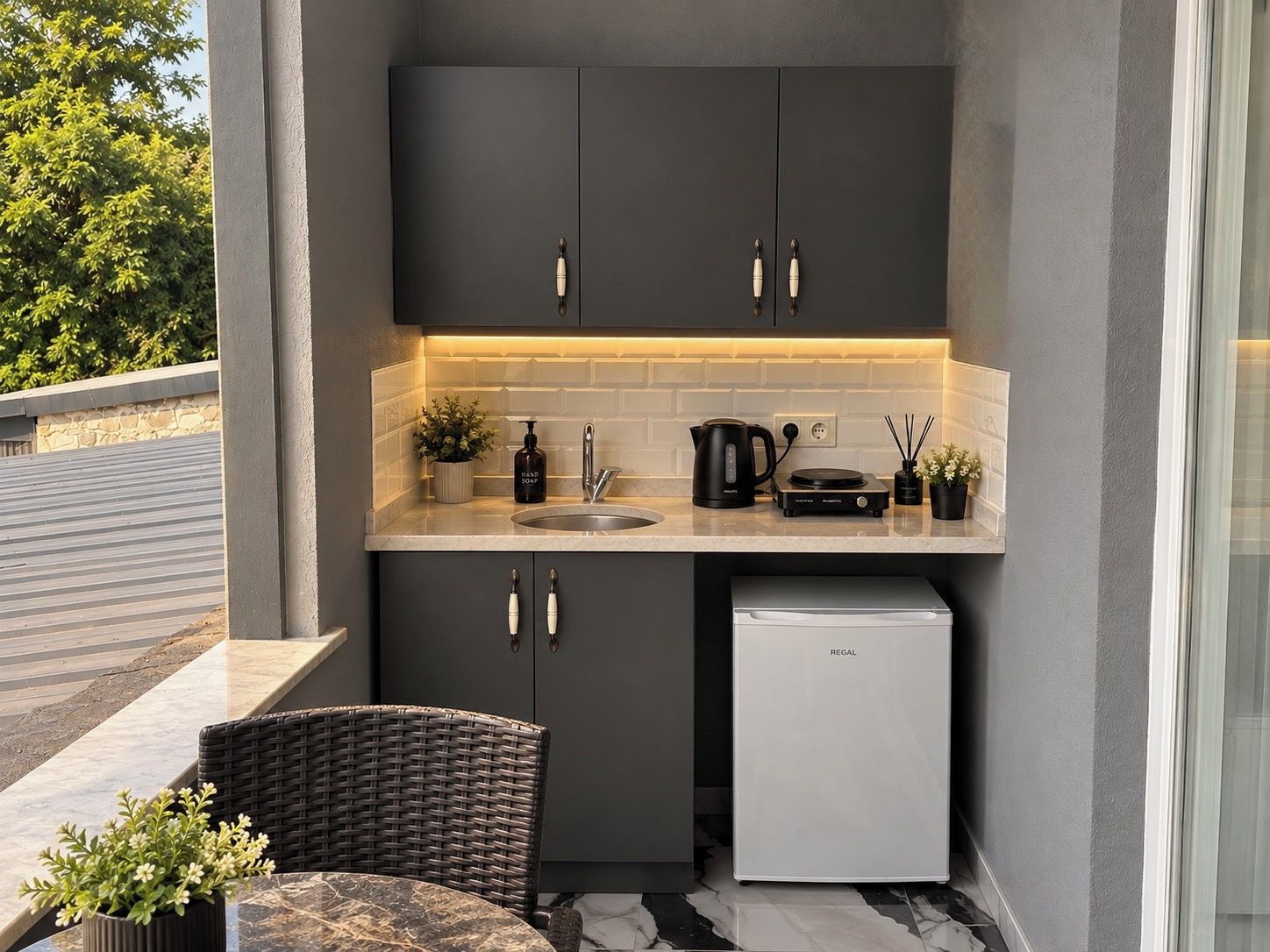 Una cocina compacta con armarios oscuros, un lavamanos, una cafetera y un refrigerador pequeño.