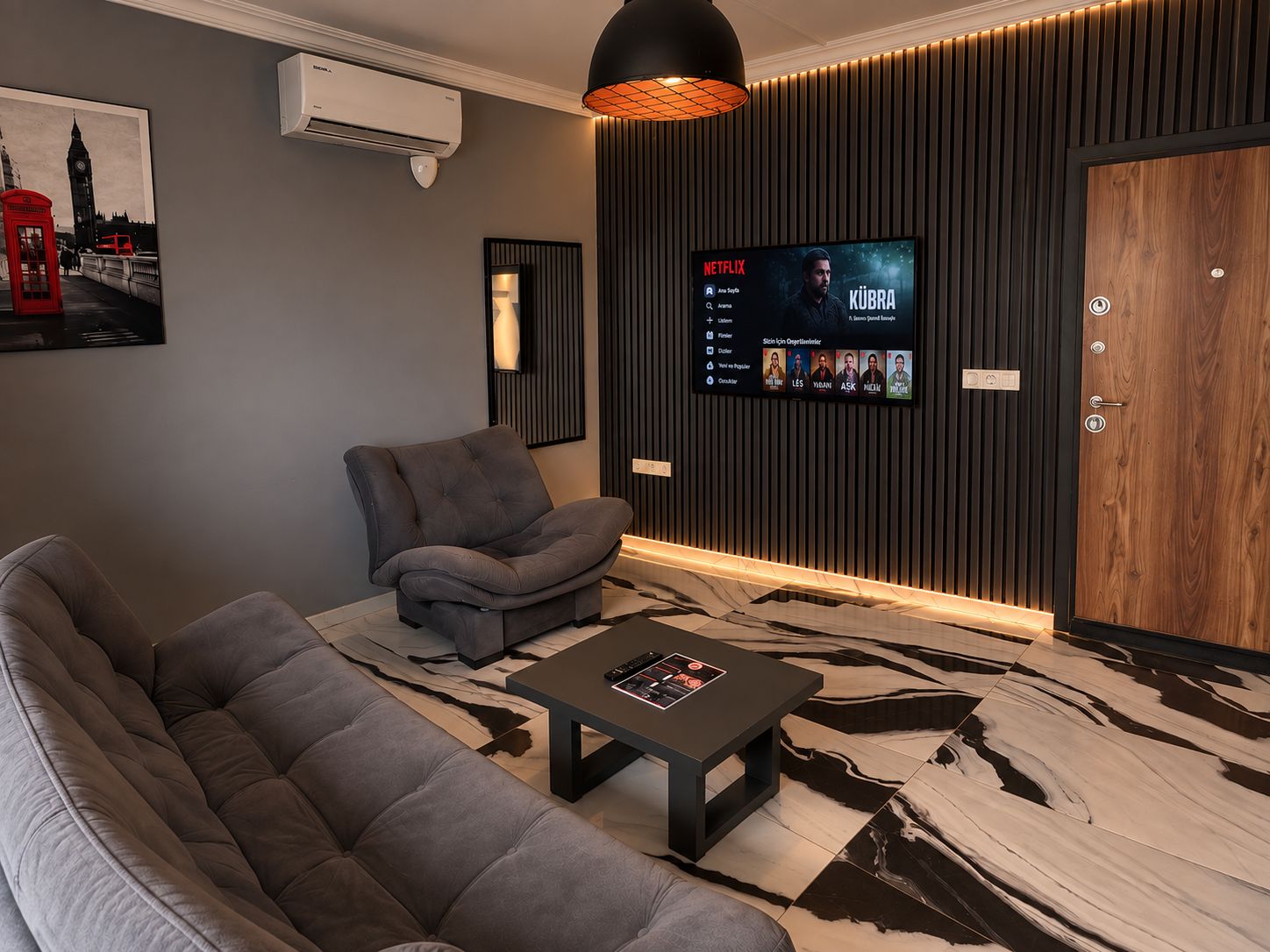 A modern living room with a sofa, armchair, and a coffee table. A flat-screen TV mounted on the wall displays a movie.