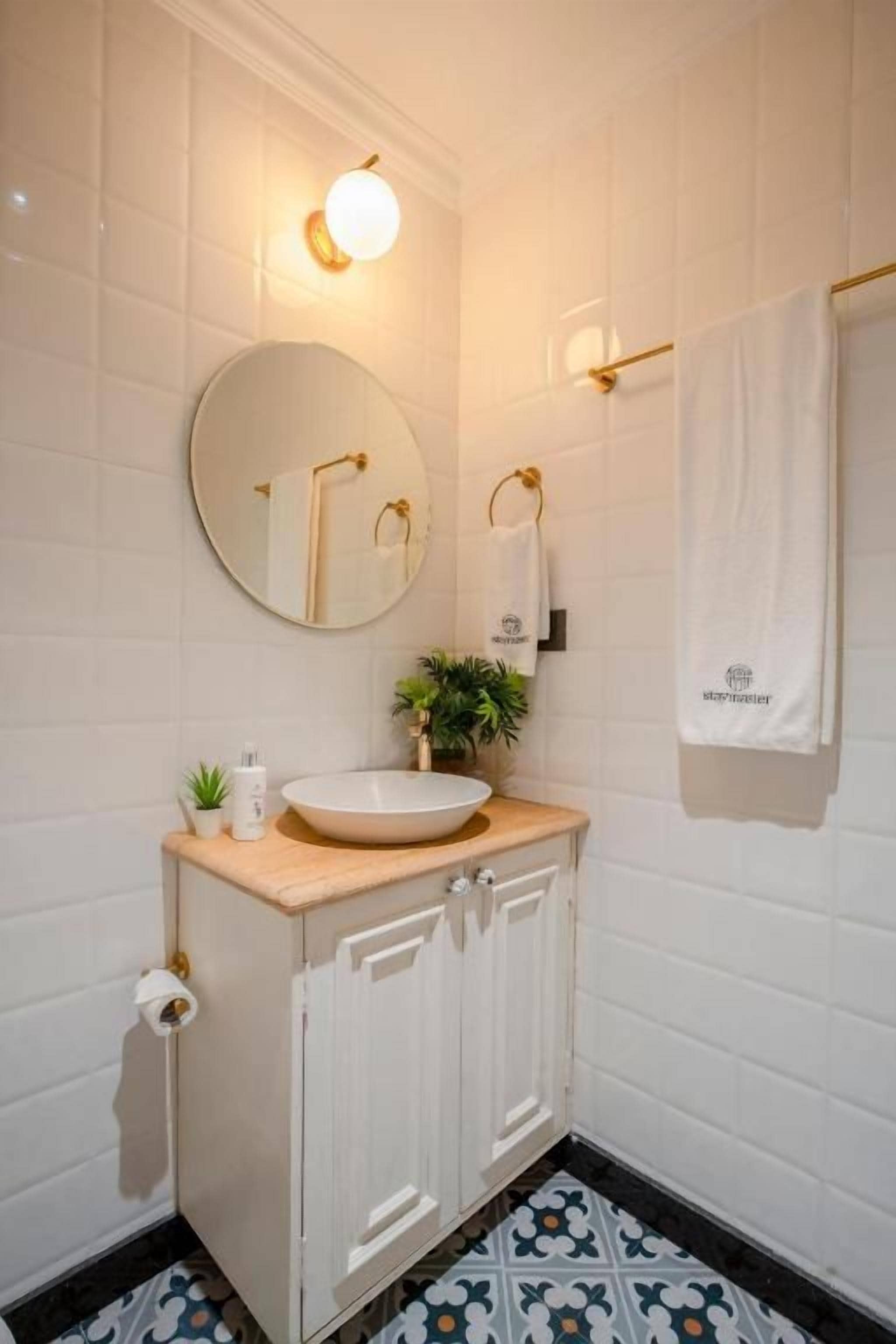 Une salle de bain avec un miroir rond, un lavabo doté d’un plan de travail en bois, un porte-serviettes et un carrelage à motifs.