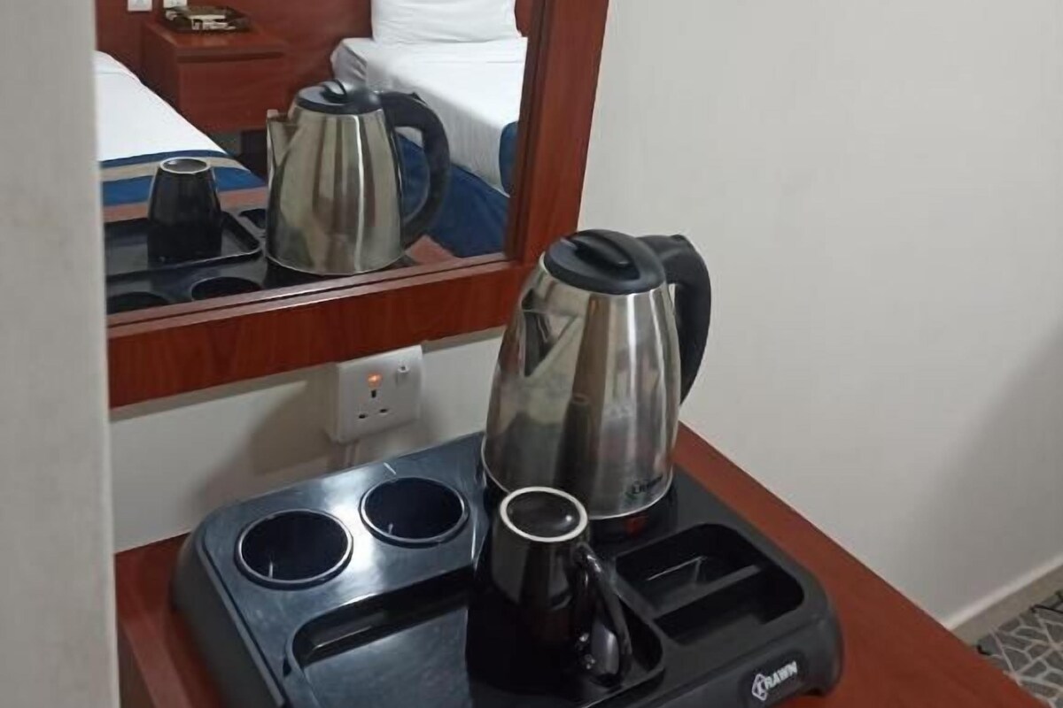 Ein Hotelzimmer mit einem hölzernen Kopfteil, einem Bett mit weißer Bettwäsche, einem Nachttisch mit Telefon, einer Kaffeemaschine und einem Getränkehalter.
