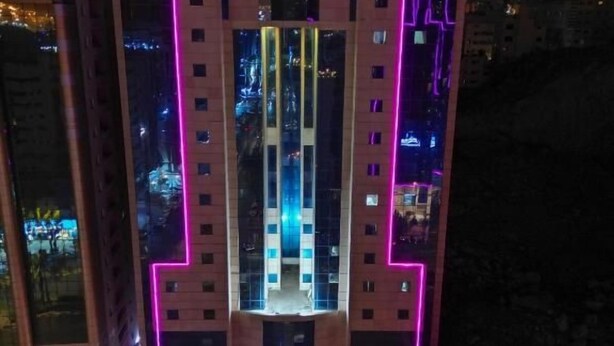 Um edifício com várias andares e fachada iluminada por neon, área de estacionamento com vários carros e ponto de ônibus.