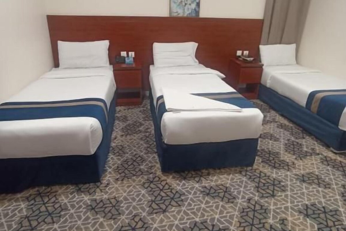 Ein Hotelzimmer mit drei nebeneinanderliegenden Betten, einem Holz-Kopfteil, einem gemusterten Teppich und einem Bild an der Wand.