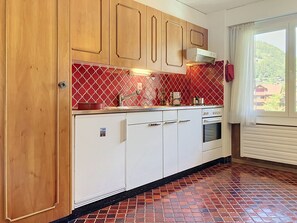 A kitchen with wooden cabinets, a red patterned backsplash, and a window with a view of a hillside. - Grand-Pré B by Interhome (Champery)