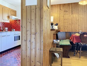 A room with a wooden wall, a kitchen area with white cabinets, and a dining table with chairs. - Grand-Pré B by Interhome (Champery)