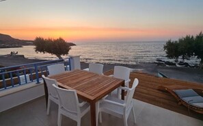 Un espace extérieur aménagé avec une table en bois et des chaises blanches, offrant une vue sur une étendue d’eau au coucher du soleil.