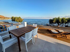 Une terrasse avec une table en bois et des chaises en osier blanc, donnant sur une plage et la mer.