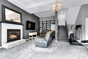 A living room with a fireplace, a mounted TV, a leather sofa, a bookshelf, and a staircase.