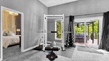 Et værelse med en seng, et træningsområde med en squat-stander og vægte, samt udsigt til en have gennem et vindue.
