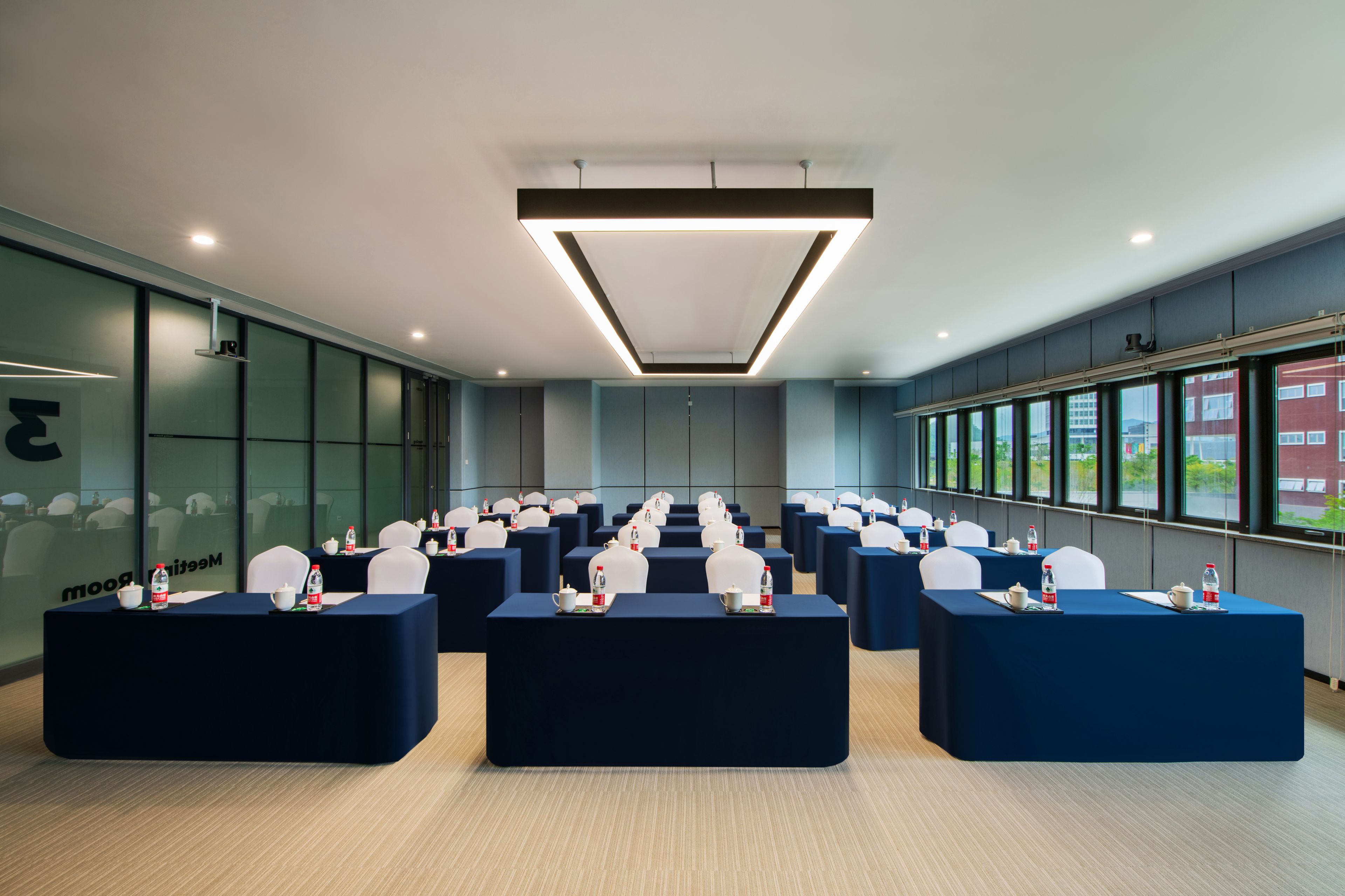 Un salón de conferencias con filas de mesas, sillas y botellas de agua, con un diseño moderno y amplias ventanas.