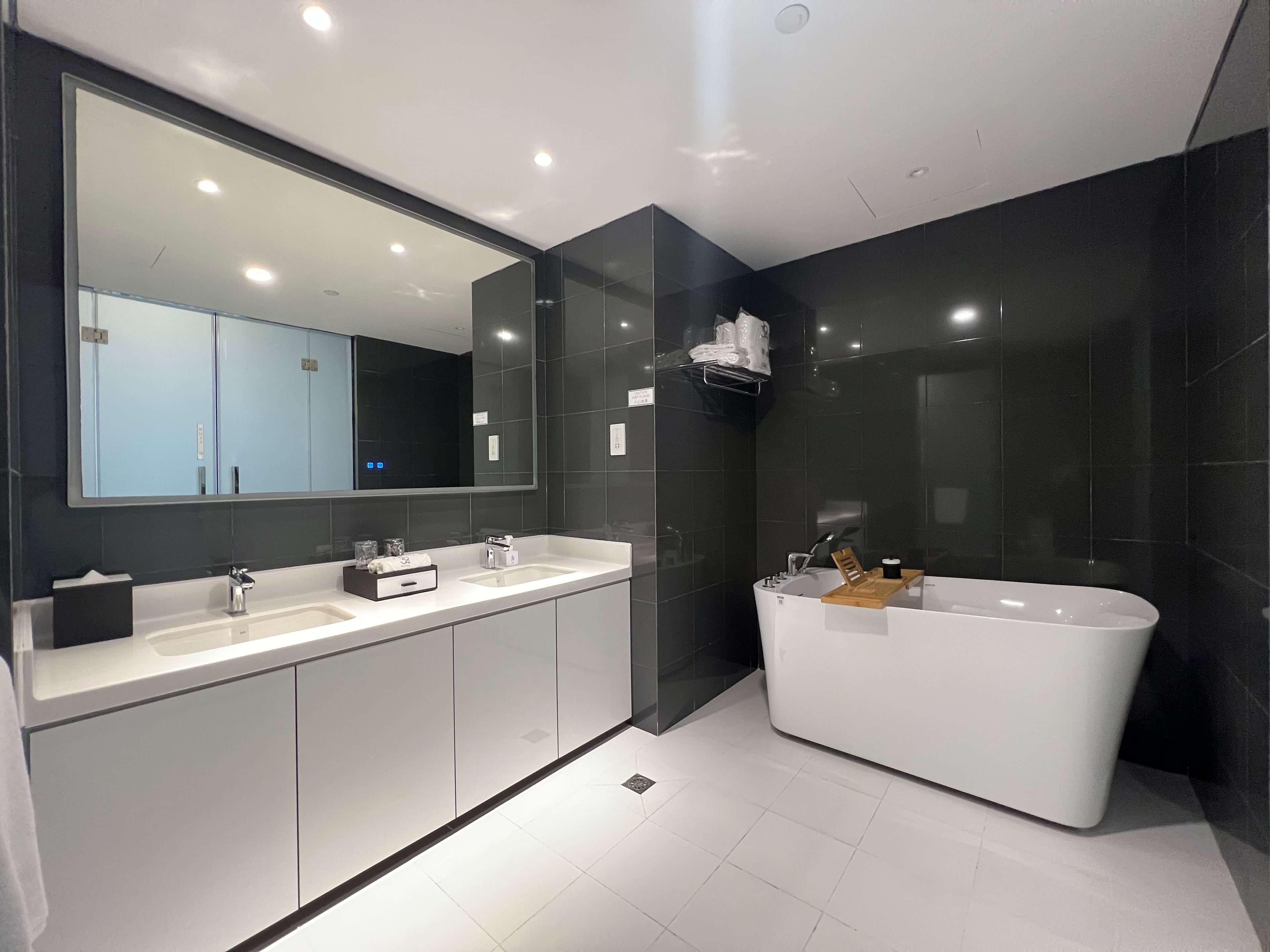 A modern bathroom with a large mirror, two sinks, a bathtub, and dark tiled walls.