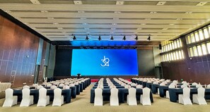 A conference room with rows of tables and chairs, a large screen displaying the number 34, and a stage area.