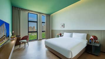 A hotel room with a large bed, a desk with a chair, a nightstand, and a view of the cityscape through the windows.