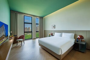 A hotel room with a large bed, a desk with a chair, a nightstand, and a view of the cityscape through the windows.