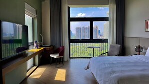 Habitación de hotel con cama, escritorio con silla, televisión y balcón con vista a edificios altos.