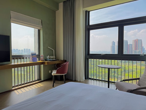 A hotel room with a bed, desk, chair, and large windows offering a city view.