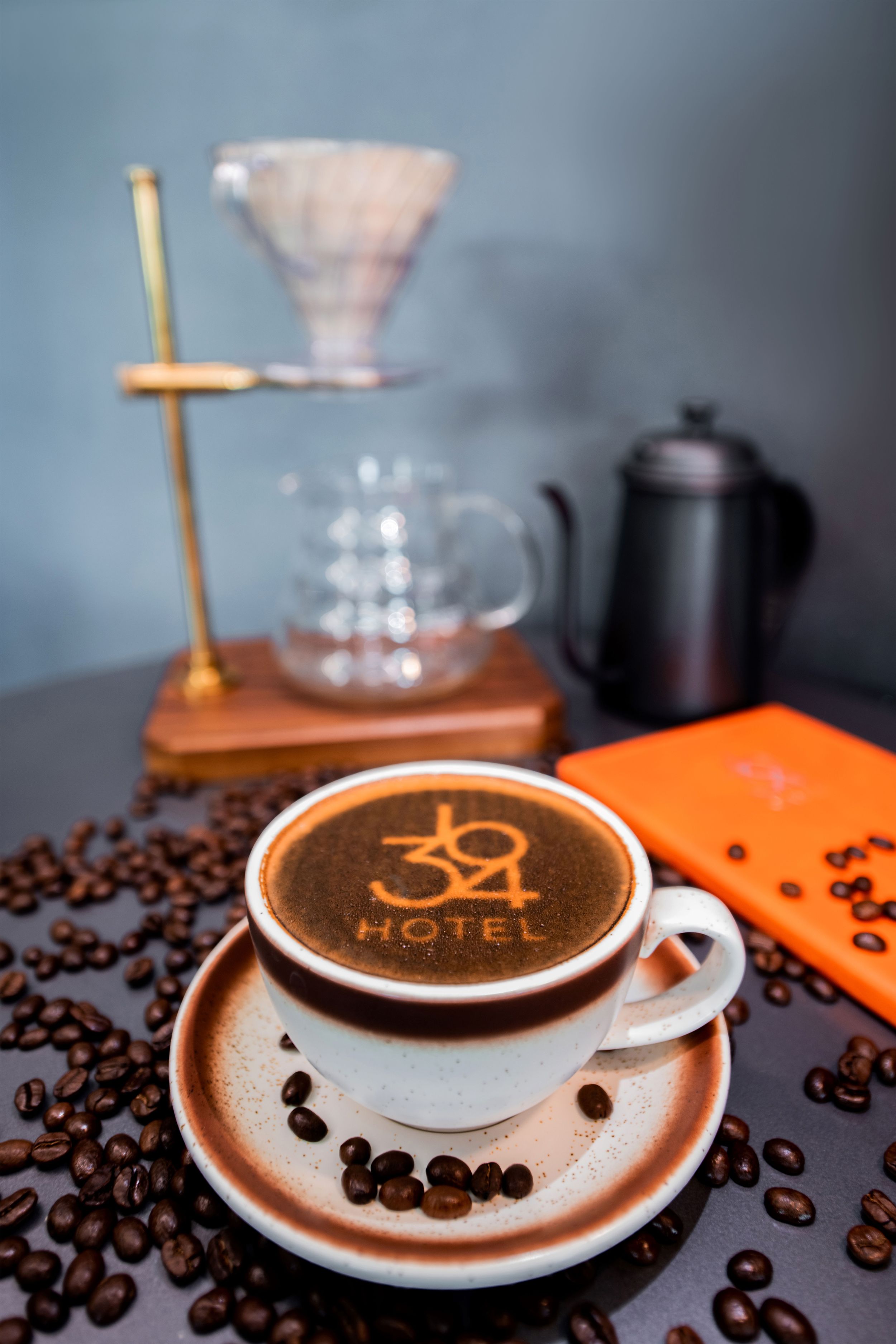 En kop kaffe med et logo i skummet, omgivet af kaffebønner på en mørk overflade.